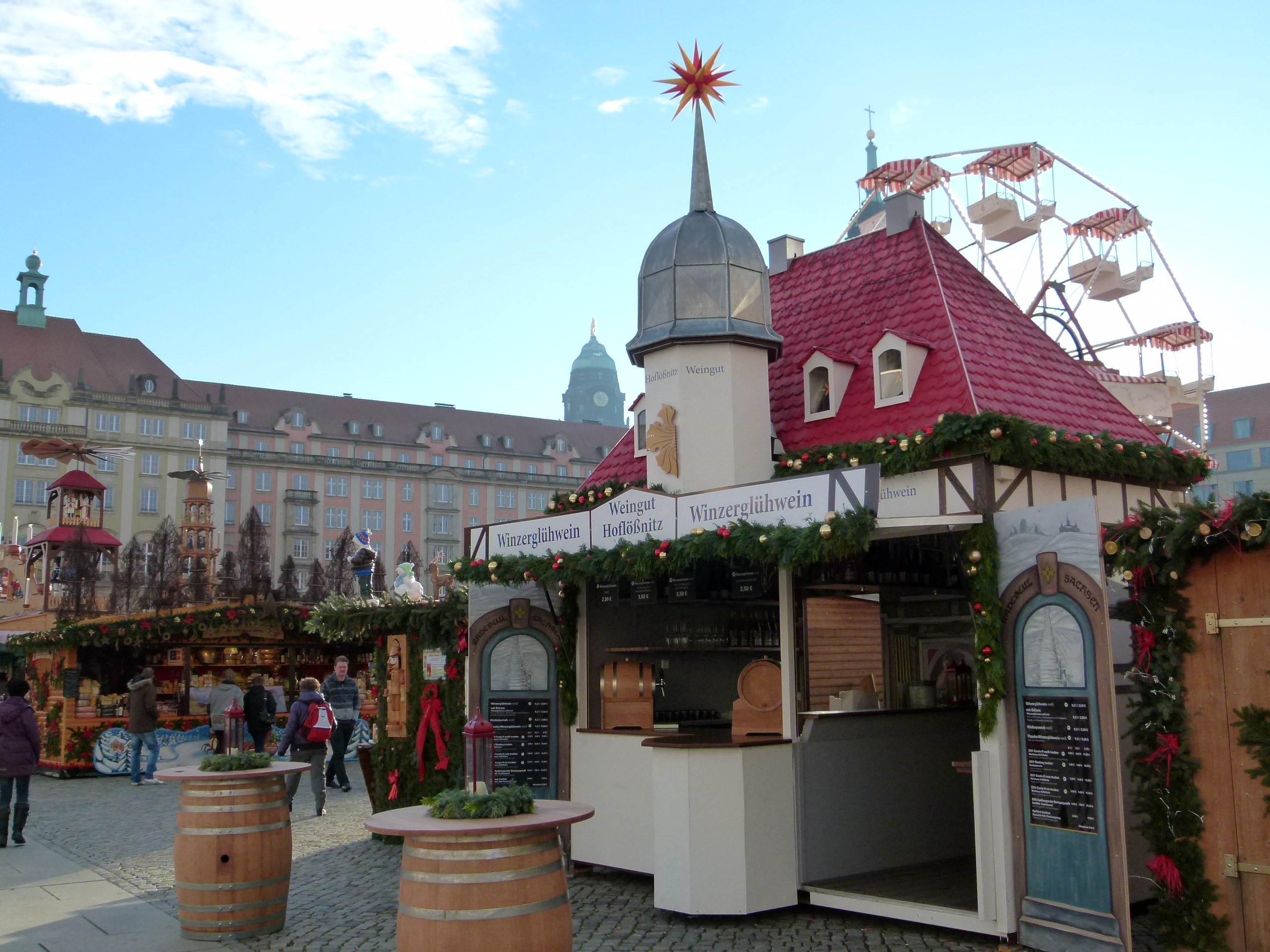 Ein geschäftiger Weihnachtsmarkt in Nürnberg, Deutschland, mit Menschen um geschmückte Stände, festliche Lichter und Schmuck, Gebäuden im Hintergrund, einem Riesenrad und einer Tafel auf der rechten Seite.
