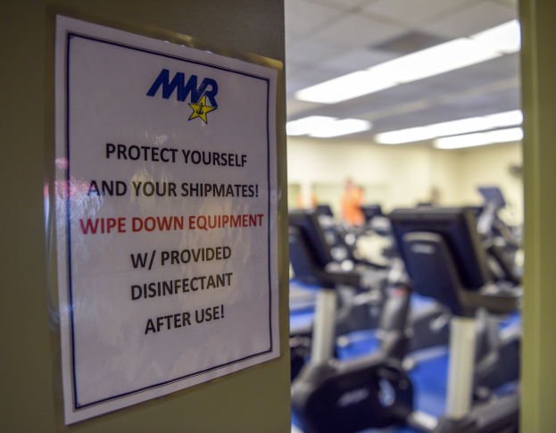 Schriftzug an einer Turnhallentür mit der Aufschrift "Schützen Sie sich und Ihre Schiffskameraden" mit Laufbändern und Deckenleuchten im Hintergrund.