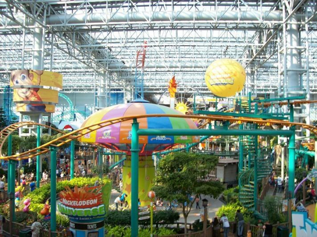 Großer Indoor-Wasserpark mit Achterbahn, umgeben von Menschen, Bäumen, Pflanzen und deckenmontierten Stangen und Lichtern, die eine lebendige Atmosphäre schaffen.