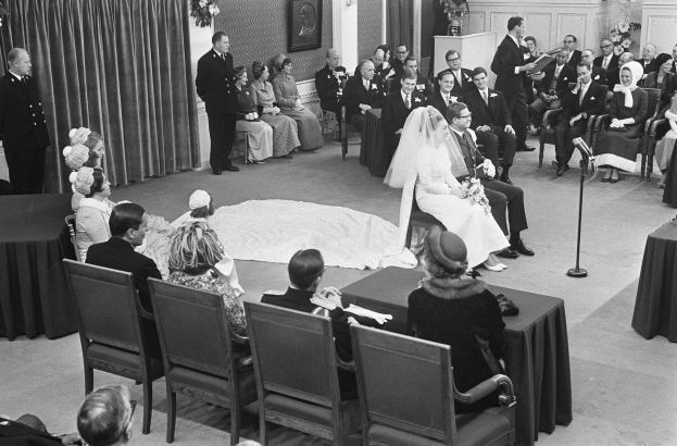 Schwarzes und weißes Foto einer Hochzeitszeremonie mit sitzenden und stehenden Menschen, einem Mann mit Mikrofon, einer Frau mit einem Strauß, einem Podium mit Mikrofon, Blumenschmuck, einem Vorhang, einem gerahmten Foto an der Wand und einer Tür im Hintergrund.