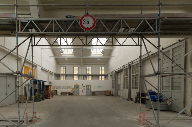 Ein großes Lagerhaus mit umfangreichen Gerüsten, Verkehrsleitkegeln auf dem Boden, Metallstangen, ein Schild, verschiedene Maschinen, eine Tür, Fenster, eine Wand, ein Dach mit Deckenleuchten.