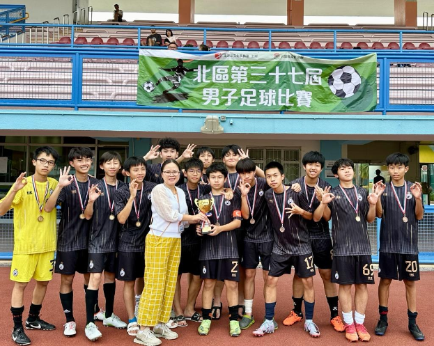 Gruppe junger Männer in Fußballtrikots auf einem Feld stehend, Medaillen tragend und einen Pokal haltend, mit einem 'Yokohama U-16 Boys Soccer Team'-Banner im Hintergrund und Zuschauern in der Nähe.