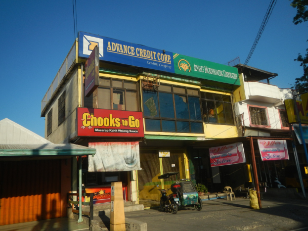 Ein Gebäude mit einer Tafel, auf der 'Advance Credit Corp' steht, an einer Straßenecke umgeben von Fahrzeugen, Strommasten und Bäumen unter einem klaren Himmel.