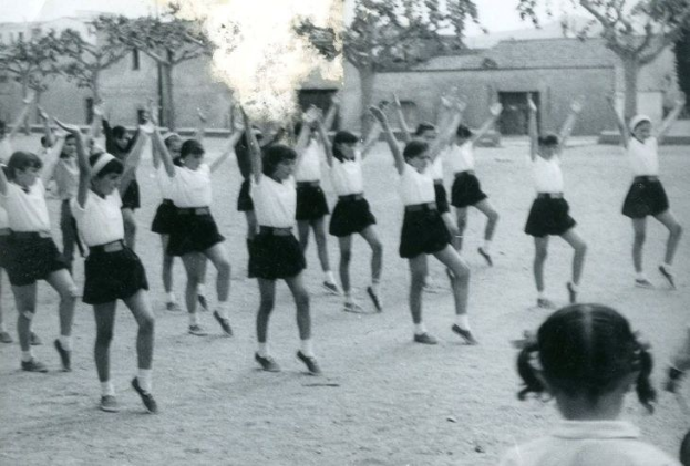 Schwarzes und weißes Foto von jungen Mädchen in Schuluniformen, die auf einer von Gebäuden und Bäumen gesäumten Straße tanzen, mit einer Person in Schuhen am unteren Bildrand.