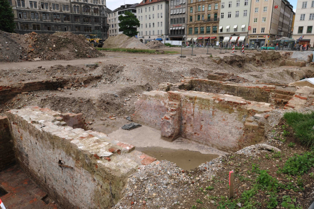 Römische Stadtruinen umgeben von modernen städtischen Elementen, einschließlich Gebäuden, Bäumen, Fahrzeugen und Menschen, mit Erde und Gras auf dem Boden.