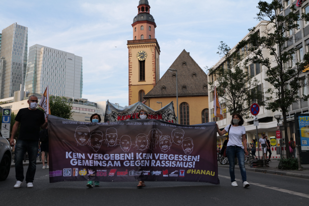 Eine Gruppe von Menschen mit Masken, die eine Straße entlanggehen und ein Banner halten, mit einem geparkten Auto auf der linken Seite, Gebäuden und Bäumen im Hintergrund und einem Uhrenturm unter einem klaren blauen Himmel.
