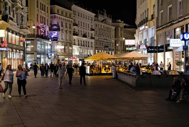 Eine belebte Stadtstraße bei Nacht mit Menschen, die gehen, auf Bänken sitzen und stehen, umgeben von Gebäuden mit Fenstern, Laternen und Texttafeln, mit Zelten und anderen Gegenständen im Hintergrund und dem Himmel darüber.