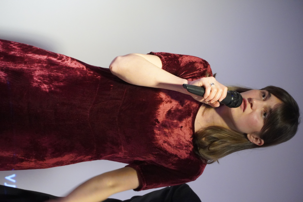 Eine Frau in einem roten Kleid steht selbstbewusst auf einer Bühne, hält ein Mikrofon in der Hand und hat einen eleganten Bob-Haarschnitt und natürliches Make-up.