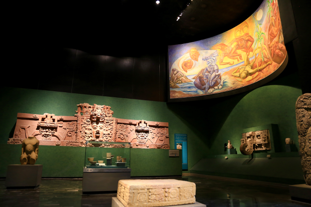 Ein Raum im Mexiko-Stadt-Museum der Anthropologie voller verschiedener Kunstwerke, darunter Skulpturen, Objekte auf Tischen, eine Texttafel, eine Wandmalerei und Deckenlampen.