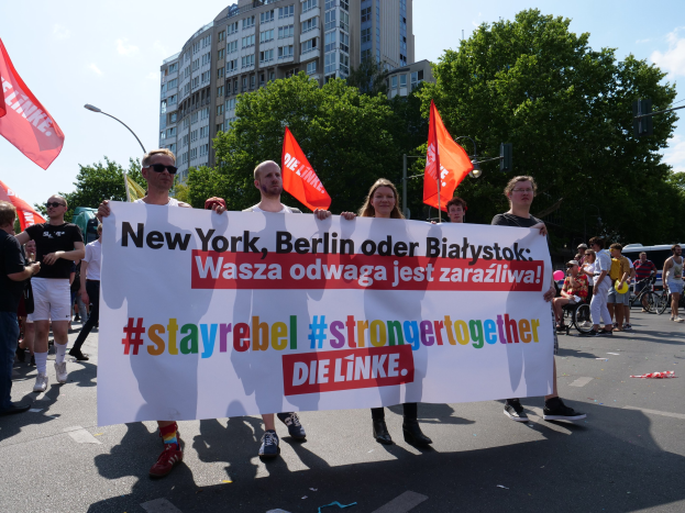 Eine Gruppe von Menschen geht eine von Bäumen gesäumte Straße in Berlin, Deutschland entlang, die ein Banner trägt, auf dem "Stay Rebel Stronger Together" steht, mit Gebäuden und Laternenmasten im Hintergrund und einem bewölkten Himmel.