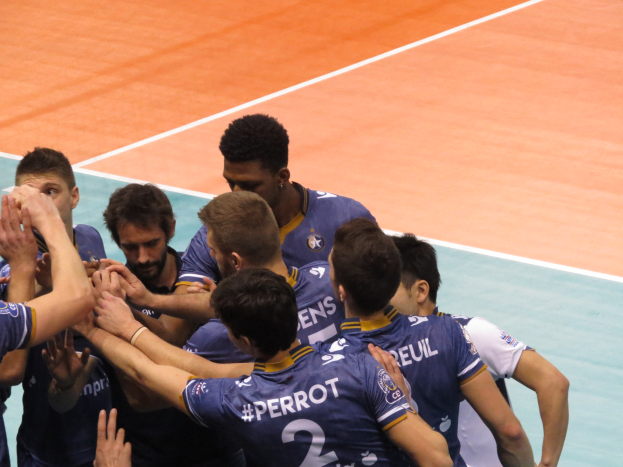 Eine Gruppe von Männern in blauen 'Ligue 1'-T-Shirts feiert auf einem Volleyballfeld, einige halten die Hände siegreich in die Höhe.