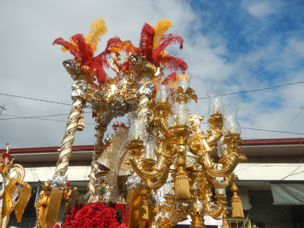 Ein großes goldenes und rotes Festwagen-Schmuck mit Blumen und Verzierungen bei einem Karnevalsumzug, mit einem Gebäude, Strommasten mit Drähten und einem bewölkten Himmel im Hintergrund.