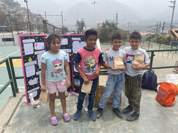 Gruppe von Kindern auf einem Basketballfeld, die Papiertüten halten, mit Tüten und Gegenständen rechts und einer Tafel mit Text links, vor einem Hintergrund aus Zaun, Pfosten, Laternen, Strommasten, Bäumen, Gebäuden, Hügeln und Himmel.