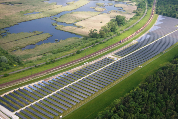 Luftaufnahme eines Solarparks mit Solarmodulen, umgeben von Bäumen, Gras, Pflanzen und Wasser, mit einem Zug auf einer nahen Bahnstrecke.