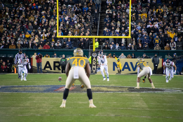 Ein Fußballspiel wird in einem Stadion mit Spielern in Sportbekleidung und Helmen im Vordergrund und einer Zuschauermenge im Hintergrund sowie sichtbaren Pfählen und Tafeln mit Text gespielt.