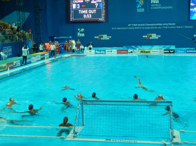 Eine Gruppe von Menschen, die Wasserball in einem Pool mit einem Netz spielen, umgeben von Zuschauern, Tafeln mit Text und Logos, Trägern und einem Bildschirm mit Zahlen und Text bei den FINA-Weltmeisterschaften 2015 in Kazan.