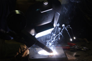 Ein Schweißer arbeitet an einem Metallstück in einer dämmrig beleuchteten Werkstatt, erhellt von einem Schweißbrenner, mit Werkzeugen auf einem nahen Tisch verstreut.