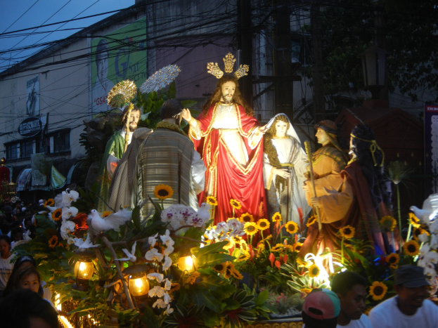 Gruppe von Menschen, die um einen Paradewagen mit Statuen von Jesus, Blumen, Blättern und Lichtern stehen, mit Gebäuden, Bäumen, Polen, Drähten und einem klaren blauen Himmel im Hintergrund.