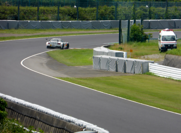 Ein Rennwagen fährt eine Rennstrecke hinunter mit einem Polizeiwagen im Hintergrund, umgeben von Gras, Pflanzen, Bäumen und Zaun, mit einem Fahrzeug auf der rechten Seite und Text unten.