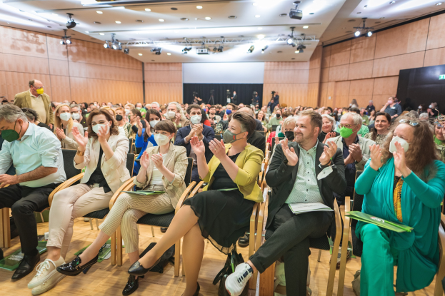 Eine Gruppe von Menschen in Stühlen klappend, einige mit Masken, mit Taschen auf dem Boden, vor einer Menge bei einer Konferenz mit einem Bildschirm und Deckenleuchten.