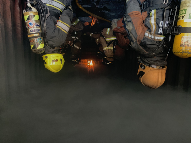 Feuerwehrleute in Schutzanzügen und Atemschutzgeräten arbeiten in einem raucherfüllten Tunnel, mit einer Lichtquelle im Hintergrund.