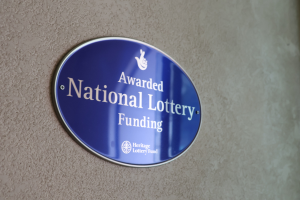 Ein rechteckiges blaues Schild mit weißem Hintergrund und schwarzer fetter Schrift in der Mitte, das "Awarded National Lottery Funding" lautet.