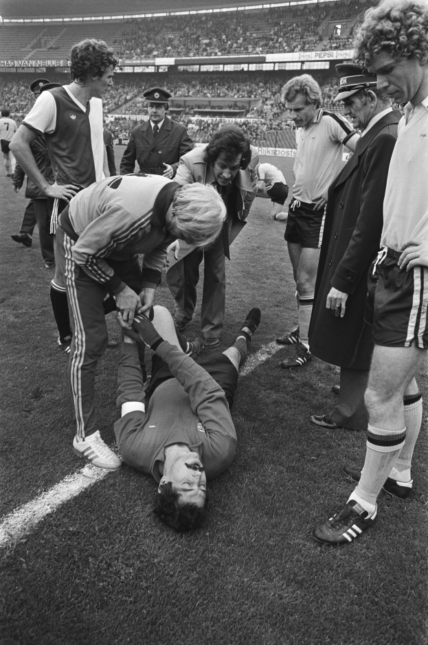 Schwarzes und weißes Foto eines Fußballspielers, der auf dem Boden liegt und von einer Gruppe Männer in einem Stadion umgeben ist, mit einer Zuschauermenge im Hintergrund.