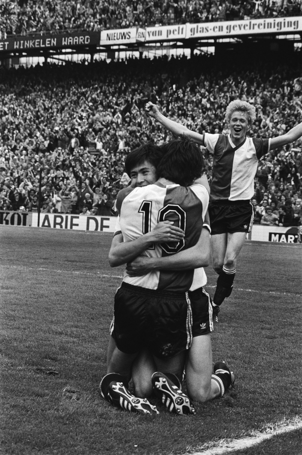 Drei Fußballspieler feiern auf dem Boden nach einem Torerfolg, mit jubelnder Menge und schwenkenden Fahnen in einem schwarz-weißen Stadion.