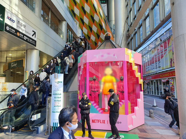 Eine Gruppe von Menschen steht vor einem Gebäude mit Glaswänden, Säulen und Texttafeln, einige tragen Masken, mit einer Rolltreppe im Blick; eine pinkfarbene Box in der Mitte zeigt Schaufensterpuppen und trägt "Netflix's new Netflix booth in Tokyo."