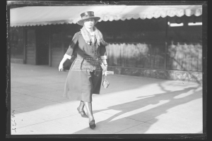 Eine stilvolle Frau in einem Kleid und Hut geht die Straße entlang in Schwarz-Weiß, trägt eine Handtasche und hohe Absätze.