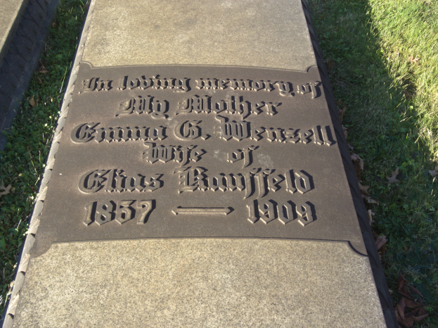Grabstein mit Inschrift "Die liebevolle Erinnerung an meine Mutter" in einem grasbewachsenen Friedhof mit trockenen Blättern.
