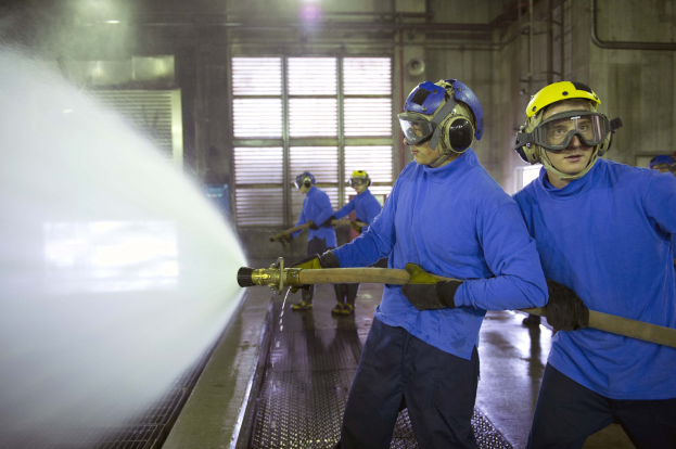 Gruppe von Männern in blauen Hemden und gelben Helmen bei der Arbeit an Maschinen, einer sprüht Wasser auf den Boden in einer Fabrik mit sichtbaren Wänden, Fenstern, Rohren und Lampen.