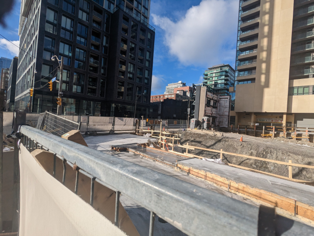 Baustelle in Toronto, Ontario mit einem großen Bauwerk im Bau, umgeben von Absperrungen, Pfählen, Verkehrszeichen und Beschilderung, unter einem bewölkten Himmel.