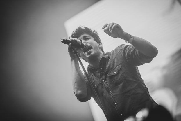 Schwarzes und weißes Foto eines Mannes, der auf der Bühne in ein Mikrofon singt, mit einem unscharfen Gesicht im Hintergrund.