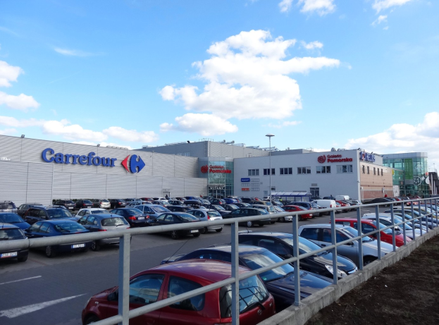 Großer Autohandel mit mehreren geparkten Fahrzeugen umgeben von städtischer Infrastruktur, einschließlich Gebäuden, Straßenlaternen und Fahrzeugen auf der Straße unter einem bewölkten Himmel.