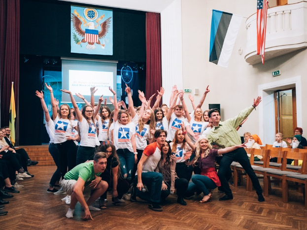 Gruppe von Menschen, die auf einem nationalen Jugendkongress-Event vor sitzenden und stehenden Zuschauern posen, mit einem Bildschirm, Flaggen und anderen eventbezogenen Elementen im Hintergrund.