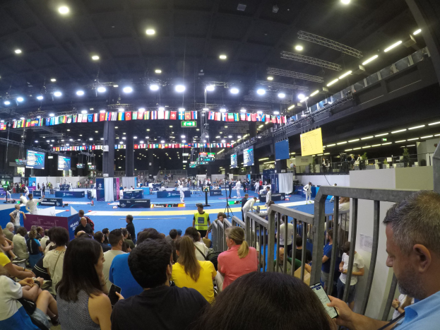 Große Menschenmenge sitzend und stehend in einer Indoor-Sportarena mit Bildschirmen, Flaggen und Deckenleuchten.