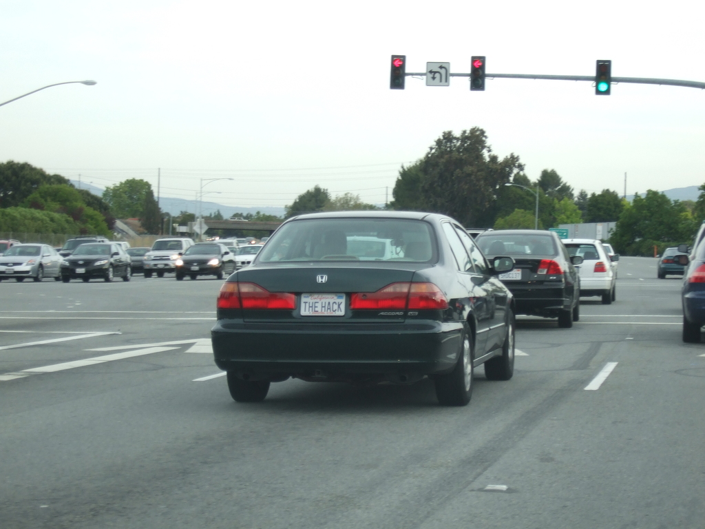Eine befahrene Straße mit mehreren Fahrzeugen, drei Strommasten und einem Verkehrszeichen auf der rechten Seite unter einem klaren Himmel.