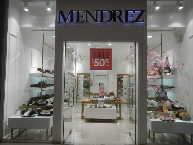 Storefront of Mendrez shoe store in Singapore with glass walls and doors, displaying shelves of shoes and bags, a text banner, ceiling lights, and visible floor.