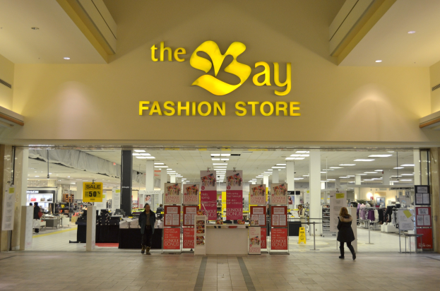 Außenansicht eines Bay Fashion-Geschäfts in einem Einkaufszentrum mit Einkäufern, Wandtexttafeln, Säulen, Deckenleuchten und Hintergrundauslagen.