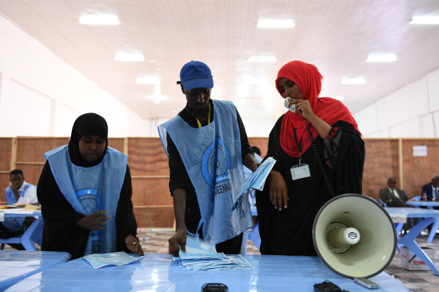 Eine Gruppe von Menschen steht um einen Tisch mit einem Megaphon, Papieren und anderen Gegenständen, mit einigen Menschen im Hintergrund auf Stühlen und Stehenden unter einer beleuchteten Decke in einer Wahllokal.