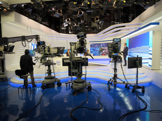 Ein Fernsehstudio mit mehreren Kameras auf dem Boden, einer sitzenden Person im Vordergrund und einigen stehenden Personen im Hintergrund, mit deckenmontierten Lampen für eine Livesendung.
