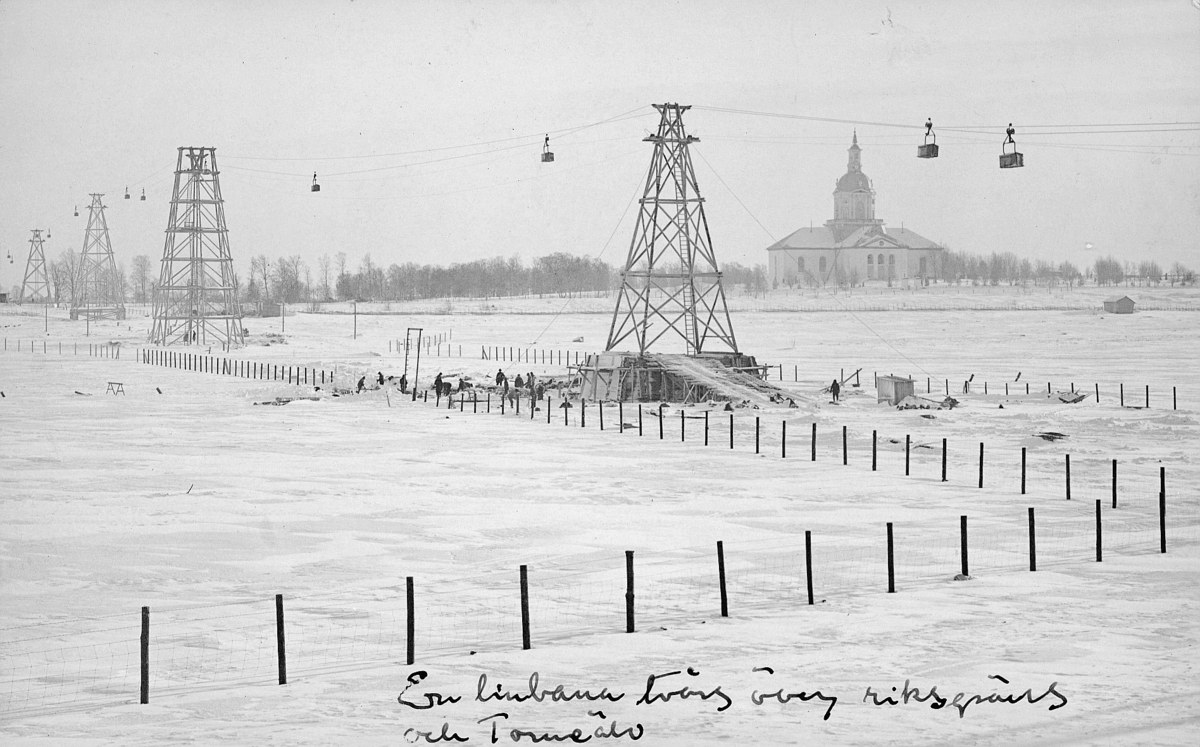 Schwarze-Weiß-Foto eines Skilifts in einer verschneiten Wiese mit Stützpfählen, Überseilbahn, Bäumen und einem Gebäude im Hintergrund, mit Text am unteren Rand.