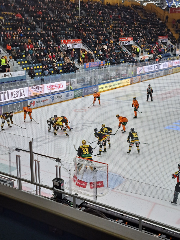 Eine Gruppe von Menschen, die Hockey auf einem Eisstadion spielen, das von einem Zaun umgeben ist, mit Zuschauern im Hintergrund und beleuchteten Stadionlampen darüber.