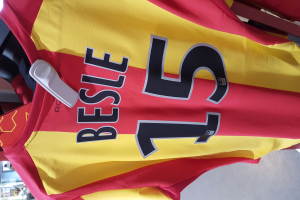 Ein rotes und gelbes Fußballtrikot mit Text und Zahlen darauf, das an einer Stange in einem Geschäft hängt, mit anderen Gegenständen und dem Boden im Hintergrund.