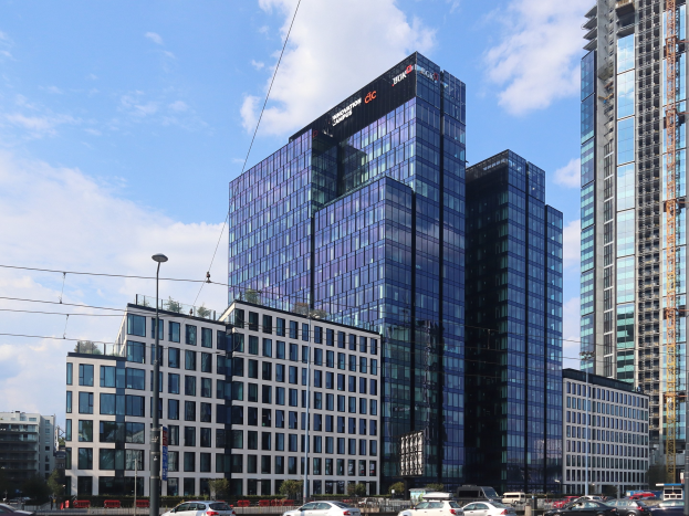 Zentrale der Deutschen Bank in Frankfurt, Deutschland, ein großes mehrstöckiges Gebäude mit zahlreichen Fenstern, umgeben von städtischer Infrastruktur einschließlich Straßenlaternen, Straßenlaternen, elektrischen Masten, elektrischen Kabeln, Kraftfahrzeugen auf der Straße, Büschen, Bäumen und einem wolkenverhangenen Himmel.