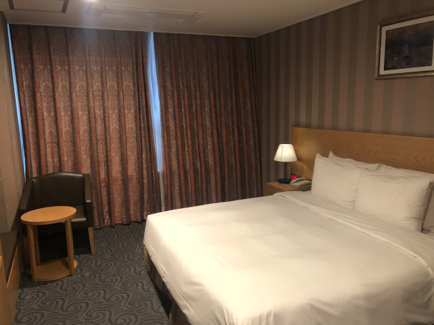 Hotelzimmer mit einem großen Bett, Stuhl, Tisch, Telefon, Lampe, Rahmen an der Wand, Fenster mit Vorhängen und Deckenbeleuchtung.