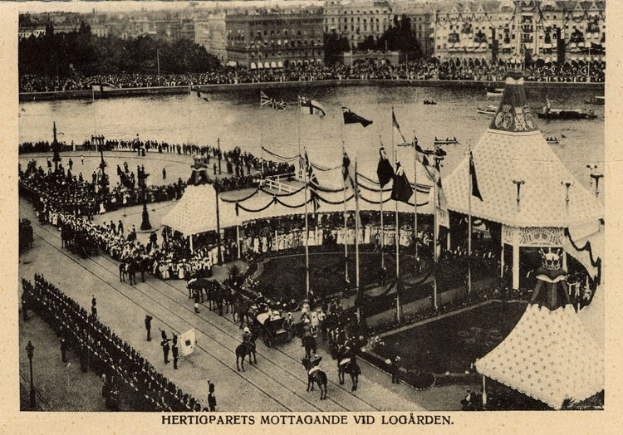 Ein altes Schwarz-Weiß-Foto einer Menge, die sich um ein Karnevalszelt mit Fahnen, Stangen und Pferden im Vordergrund und Gebäuden, Bäumen und Wasser im Hintergrund versammelt hat, mit Text am unteren Bildrand.