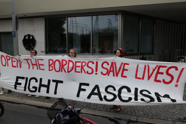 Demonstranten mit einer Banner mit der Aufschrift "öffne die Grenzen, rette Leben, bekämpfe Faschismus" vor einem Gebäude mit Glasfenstern und einem Pfahl, mit einem Fahrrad und einer Tasche auf der Straße.