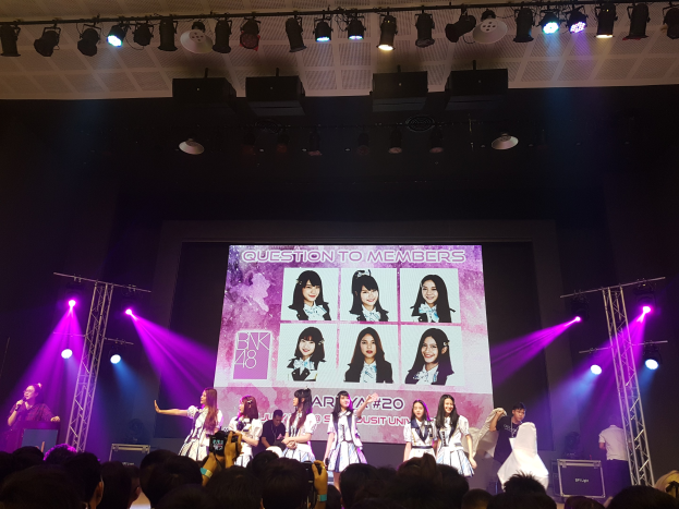 Eine Gruppe von Menschen performs auf einer hell erleuchteten Bühne mit Trägern, während eine Menge darunter das Ereignis aufzeichnet; ein großes Display im Hintergrund zeigt Bilder der K-Pop-Gruppe BK48.